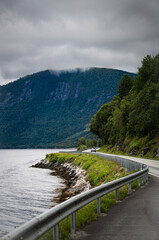 A solitary car journeys on Norwegian National Road Highway 70 beside Ålvundfjord, bordered by lush greenery and a mountain in the distance under a cloudy sky.