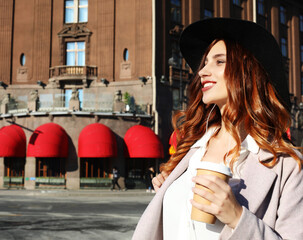 Smiling stylish young woman drinking coffee while walking on a city street