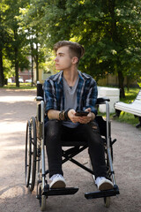 A young disabled man sits in a wheelchair and listens to music on headphones. Walk on a summer day.