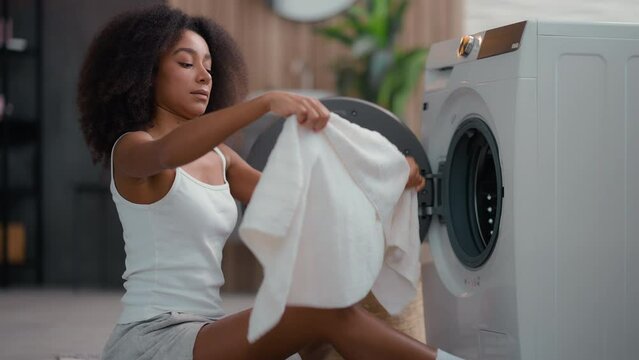 African American female woman girl ethnic lady housewife homeowner housework at floor open washing machine smell clean white towel cloth bad smelling sniff terrible aroma laundry conditioner gel stink