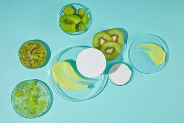 A round white podium placed above the other glass one with a yellow cream smudge decorated. Kiwi in form of slice and piece displayed on glass dishes around, photo with blue background and top view
