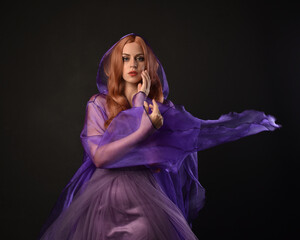 close up portrait of beautiful female model with long red hair, wearing a purple fairy  fantasy cloak with hood and gown. Elegant gestural hand poses as if casting magic spell. Isolated on dark black 