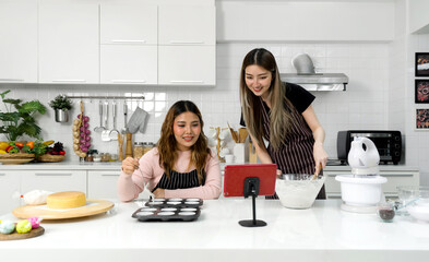 Two young women in an apron make dessert in the kitchen while looking at online recipe guide book...