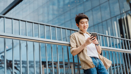 Portrait of handsome Asian student using smartphone. A young man standing outdoor happy smiling with holding mobile phone