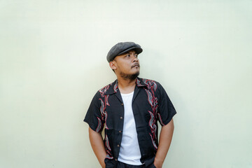 Macho man with beard and mustache looking serious isolated on beige background. Half body portrait of a vintage style Southeast Asian adult man in a patterned shirt, white tank top and newsboy cap