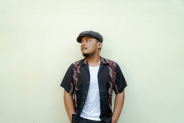 Macho man with beard and mustache looking serious isolated on beige background. Half body portrait of a vintage style Southeast Asian adult man in a patterned shirt, white tank top and newsboy cap