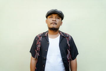 Macho man with beard and mustache looking serious isolated on beige background. Half body portrait of a vintage style Southeast Asian adult man in a patterned shirt, white tank top and newsboy cap