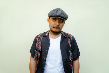 Macho man with beard and mustache looking serious isolated on beige background. Half body portrait of a vintage style Southeast Asian adult man in a patterned shirt, white tank top and newsboy cap
