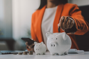 Beautiful Asian businesswoman insert coins in a pin piggy bank on the table. saving money,...