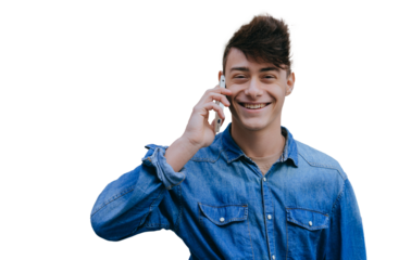 young man in a denim shirt smiles while talking on his phone, standing against transparent background