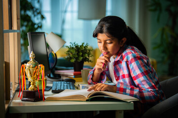 Asian Indian girl child studying at home on study table with computer, books, Globe model, victory trophy