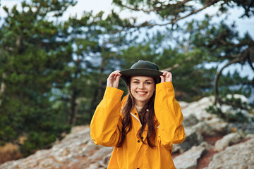 A woman in a yellow raincoat and hat enjoying a scenic view on a rocky hill with trees in the background during a hiking trip