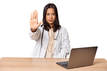 Young Filipino doctor with laptop in studio standing with outstretched hand showing stop sign, preventing you.