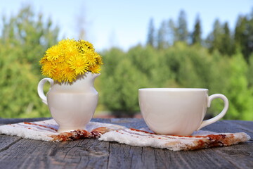 A white cup of hot tea next to a white jug with yellow flowers stands on a wooden desk on the...