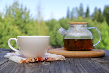 A white cup of hot tea next to a glass teapot on a tray stands on a wooden desk on the background...