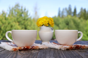 A white cups of hot tea next to a white jug with yellow flowers stand on a wooden desk on the background of summer forest landscape.