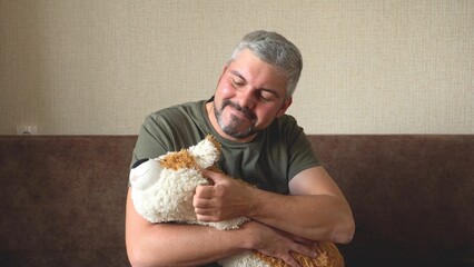 A man with a beard strokes and hugs a toy dog. Green baseball cap and T-shirt. Military. Corgi dog breed. Feelings of tenderness and love. In room. Sitting on a brown sofa 