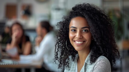 Stylish Middle Eastern Manager Sitting at Her Desk in Creative Office. Young Stylish Female with Curly Hair Looking at Camera with Big Smile. Colleagues working in the background. - Powered by Adobe