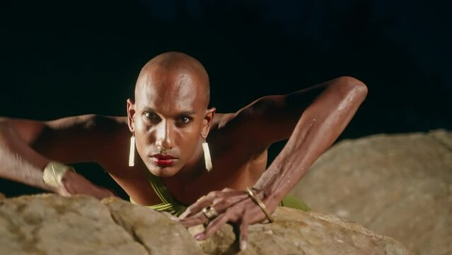 Gender fluid black person crawling like wild primeval animal on rock in creek natural water pool at night. Queer ethnic fashion model in long revealing clothes moves creepily in the middle of lake.