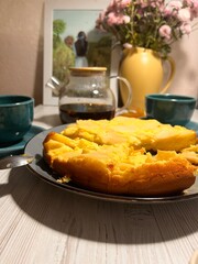 A freshly baked apple pie is on a plate in the center of the dining room table. On the table are two blue cups, a glass teapot, a yellow vase with pink flowers, and a painting