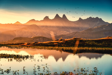 Sunrise shines over Lac Guichard with bird flying on Arves massif and lake reflection at Aiguilles...