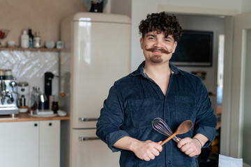 Smiling man holding wooden spoon and wire whisk in kitchen at home