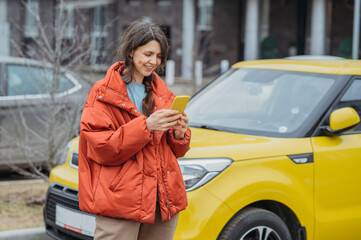 Smiling woman using smart phone standing next to yellow car at street