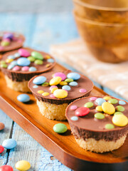 Four decorated cupcakes with chocolate coating and colorful pieces of candy lie on a wooden tray. The cupcakes rest on a light blue surface, with more candies on a blurred background.