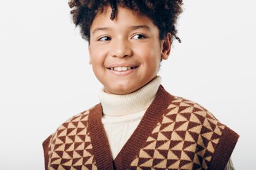 a cheerful young boy with curly hair in a brown sweater and white turtleneck smiling outdoors on a...
