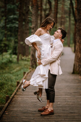 Fototapeta premium Riga, Latvia, - August 26, 2024 - Groom lifting bride in his arms on a wooden path in a forest, both smiling and looking at each other.