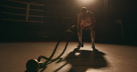 Professional boxer doing cardio or endurance training. Athletic man exercises with fighting ropes in a dark boxing gym.