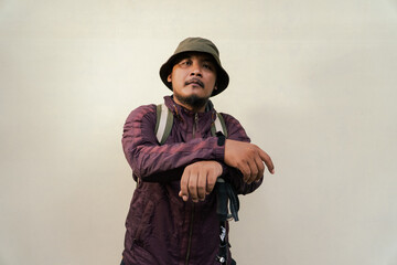 Half body portrait of adult Asian man with beard and mustache posing like an adventurer. Model in hiking clothes with jacket, bucket hat, carrier bag isolated on beige background.