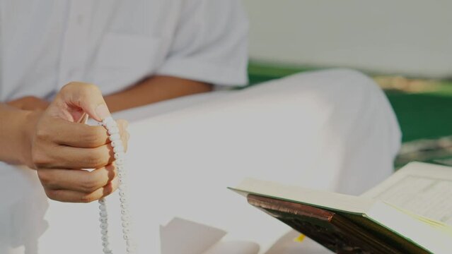 Front View Of Muslim Man Hand Holding Rosary Or Prayer Beads And Sitting Crossed Legs In Front Of Holy Quran In Mosque