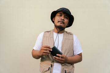 Macho asian man with beard and mustache wearing safari clothes isolated on beige background. Half body portrait of adult Southeast Asian man posing wearing vest, bucket hat and hip flask