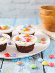 A white plate with four chocolate cupcakes topped with white icing and colorful candies. Various colorful candies are scattered on the blue wooden surface around the plate.