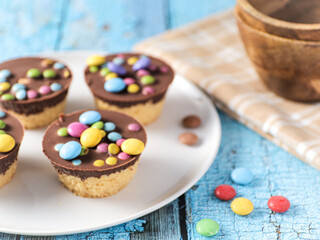 A white plate with four chocolate cupcakes topped with brown icing and colorful candies. Various colorful candies are scattered on the blue wooden surface around the plate.