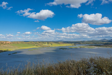 Wide landscape of the Tagus River flowing through the Burujón Canyon in the province of Toledo, Spain, with lush green fields and distant mountains.