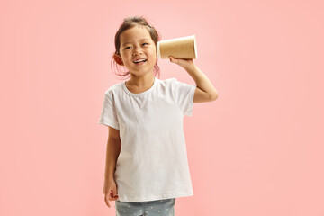 Cheerful cute Asian child girl spying using paper cup for listening, trying overheal someone, holds...