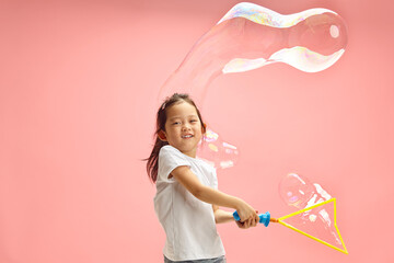 Cute Chinese Appearance Child Girl Creates Big and Long Soap Bubble on Pink Isolated Background.