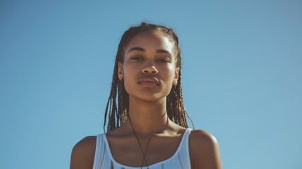 Portrait of an African black woman with long braided hair wearing a white tank top standing against a clear blue sky. looks directly at the camera with a neutral expression. - Powered by Adobe