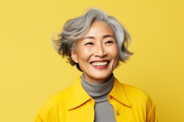 Portrait of a cheerful asian woman in her 50s smiling at the camera in front of pastel yellow...