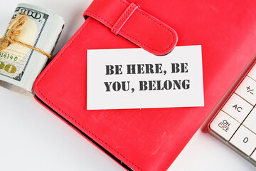 BE HERE, BE YOU, BELONG Words written on a business card on a business notebook near money and a calculator on a white background