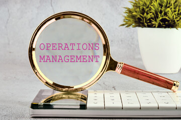 Operations management business concept. OPERATIONS MANAGEMENT written. through a magnifying glass standing on a calculator on a gray background with a plant in the background without focus