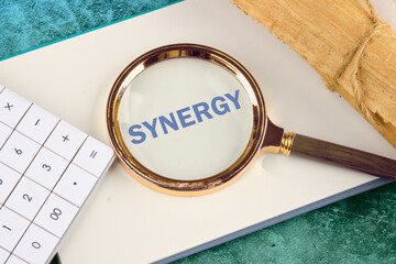 Synergy, cooperation or efficiency in business concept. The word synergy through a magnifying glass on a blank sheet of notepad next to a calculator and a roll of papyrus