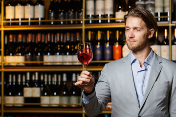 Glass of red wine on cellar background, male sommelier appreciating drink against wooden shelf with bottles.