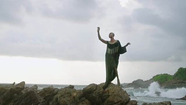 Queer black person gracefully poses in posh dress, handmade jewelry stands on scenic sea cliff top above dramatic cloudy sky, ocean waves. Lgbtq biethnic gay model in brass accessories.