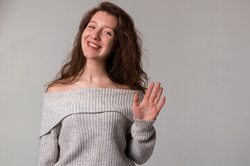 A woman with long hair and a grey sweater is happily waving her hand