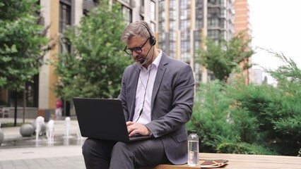 Adult businessman using headset while typing on laptop outdoors.