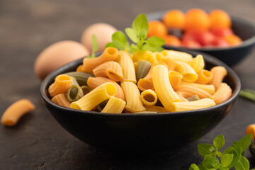 Rigatoni colored raw pasta with tomato, eggs, spices, herbs on black concrete. Side view, selective focus.