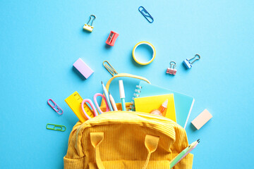 Yellow backpack with an array of colorful school supplies spilling out, including notebooks, pencils, and scissors on blue background. Back to school concept.